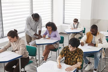 hero-img-01 A diverse group of college students taking an exam, supervised by a teacher, in a classroom setting.