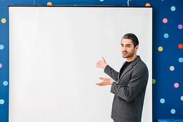 gallery-1 Man in gray blazer presenting with blank projector screen in a polka dot room indoors.