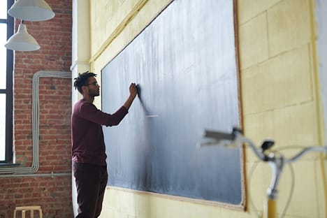 hero-img-02 Man with dreadlocks writing on a blackboard in a classroom setting.