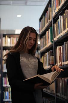 services-02 A young woman reading a book in a calm library setting, surrounded by shelves.