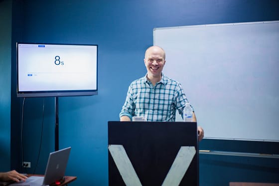about-01 Smiling man giving a lecture at a podium with a whiteboard and monitor in a classroom.